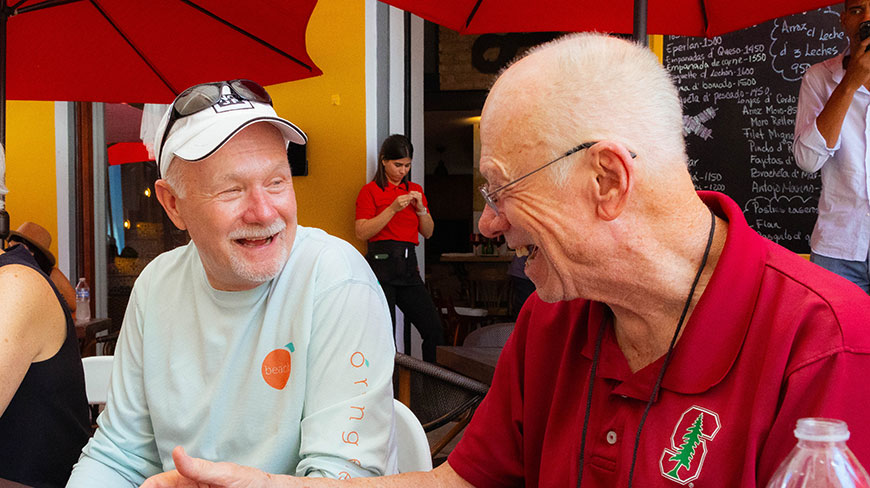 Two men laugh while sitting together at an outdoor restaurant with red umbrellas in Havana, Cuba.