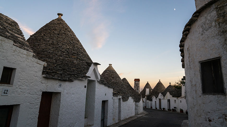 A street in Alberobello is lined with traditional whitewashed trulli houses with conical stone roofs under a twilight sky.