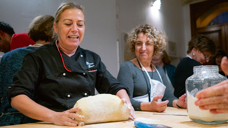 A female chef in a black jacket smiles while holding a large loaf of bread dough as another woman watches in the background.
