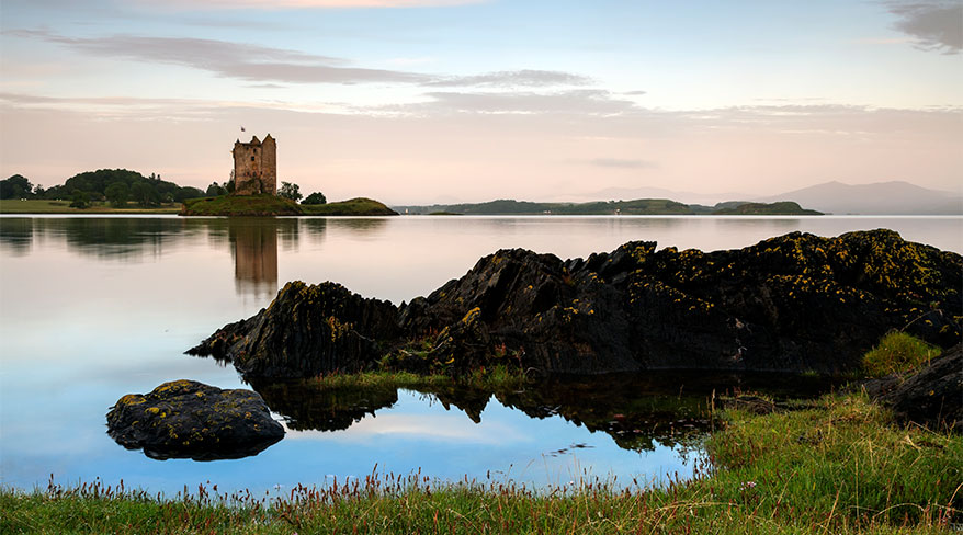 A stone castle sits on a small island in a calm loch at dusk, with dark, mossy rocks along the shoreline in the foreground.