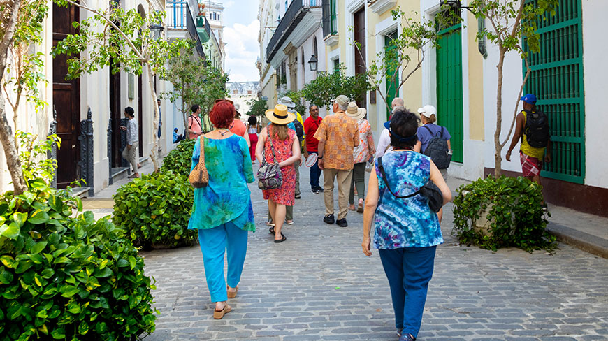 A group of people on a walking tour stroll down a cobblestone street in Havana, Cuba, lined with colonial-style buildings.