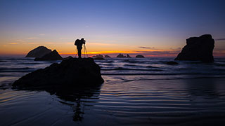 A silhouetted photographer stands on a rock at the beach setting up a tripod at sunset.