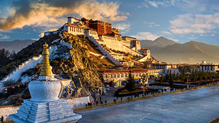The Potala Palace in Tibet at sunrise, viewed from behind a white and gold stupa with mountains in the background.