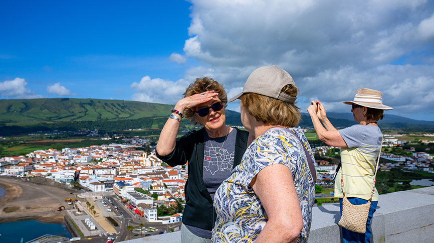 Three women stand on a scenic overlook viewing a coastal town and green hills below under a blue sky.