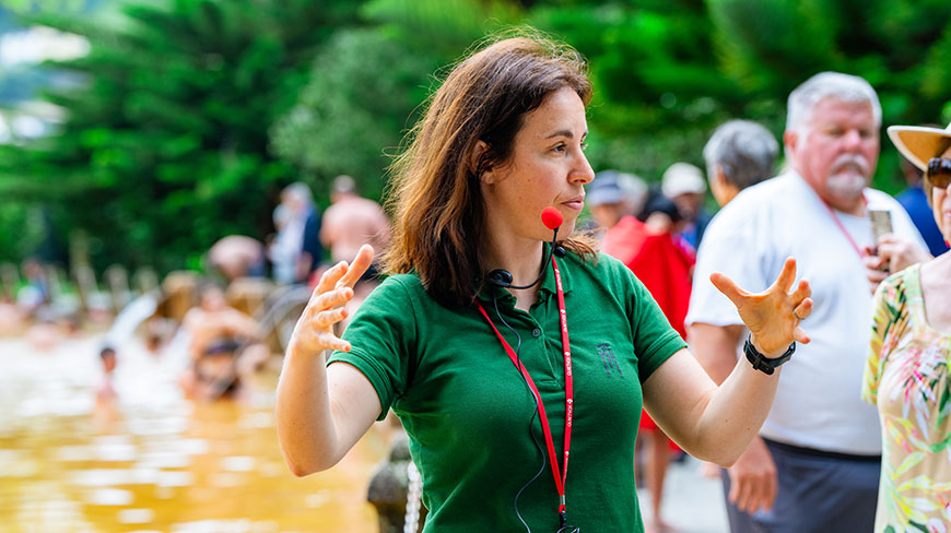 A female tour guide in a green shirt with a headset microphone speaks and gestures to a group of tourists outdoors.