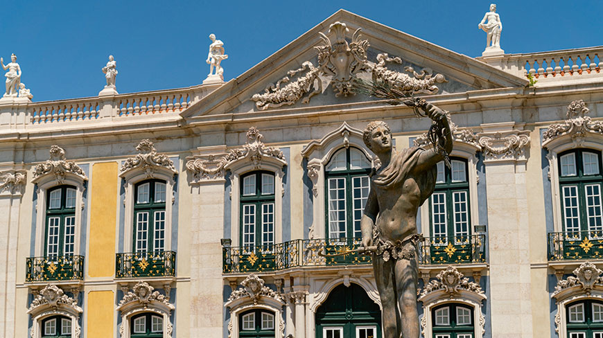 A classical statue stands before the ornate, multi-story facade of the Queluz National Palace in Portugal under a clear blue sky.