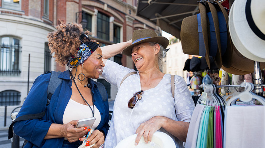 Two smiling female friends shop for hats at an outdoor market stall on a European city street.
