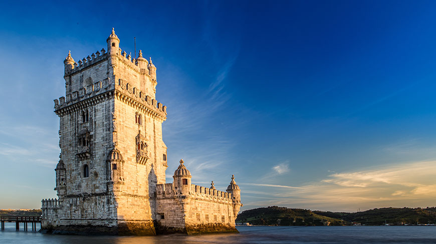 The historic Belém Tower in Lisbon, Portugal, sits on the water, illuminated by the golden light of sunset against a deep blue sky.