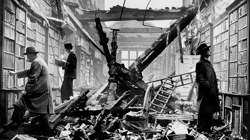 Three men in coats and hats browse books amidst the rubble of the bombed-out Holland House library in London during an air raid.