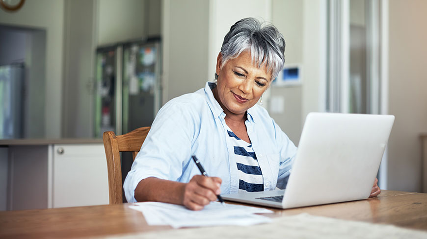A smiling woman with short gray hair writes on a paper while sitting at a wooden table with her laptop.