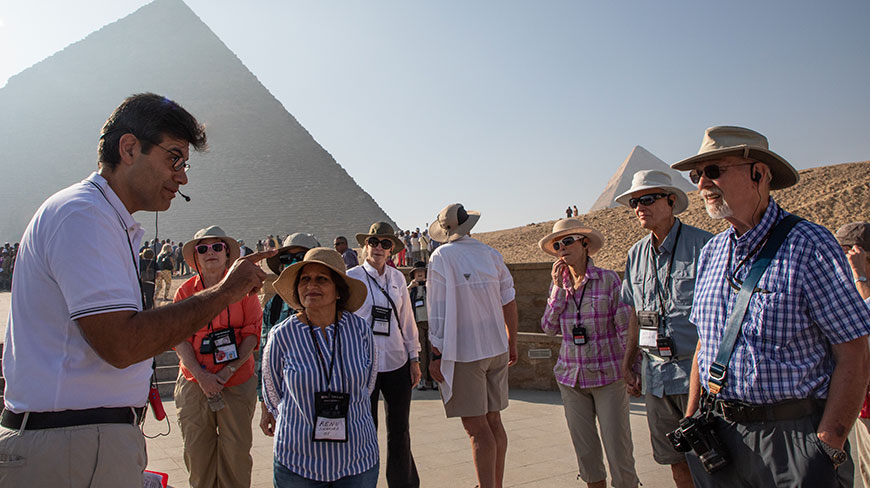 A tour guide gestures while speaking to a group of travelers with the Great Pyramids of Giza in the background.