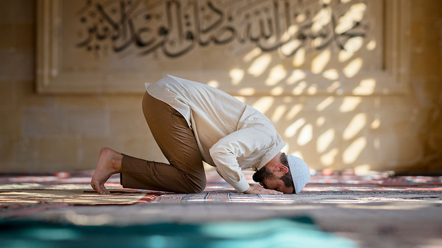 A Muslim man prostrates in prayer on a rug inside a mosque with calligraphy on the wall in the background.