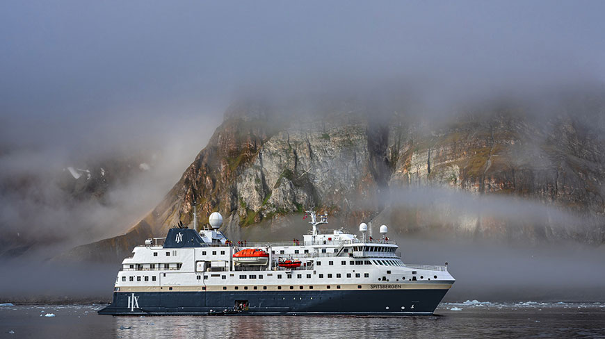 The MS Spitsbergen expedition ship sails through icy waters in front of a misty, mountainous fjord in Norway.