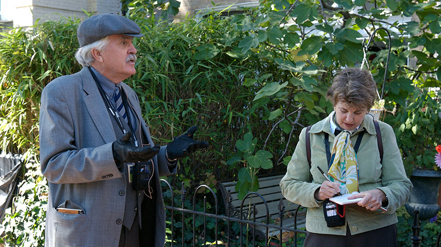 A man in a grey suit and cap leads a historical tour in Washington, D.C., as a woman takes notes.