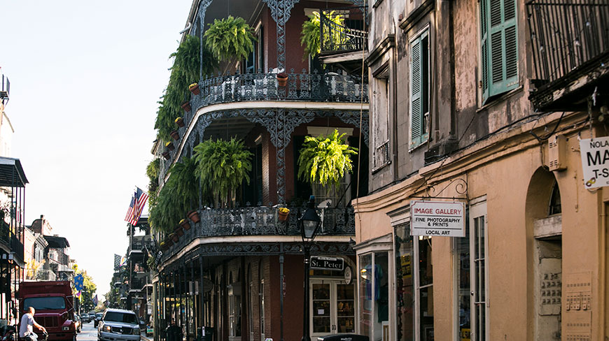 A street corner in New Orleans' French Quarter shows historic buildings with ornate, fern-draped iron balconies.