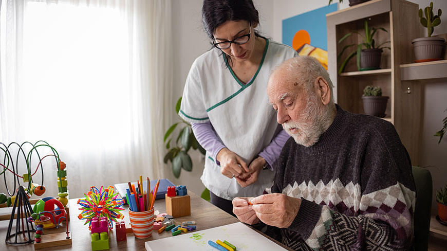 An elderly man with dementia participates in art therapy with a female caregiver.