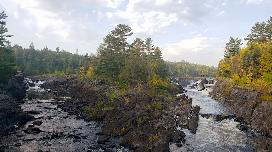 A rocky river with several waterfalls flows through a northern Wisconsin forest under a cloudy sky.