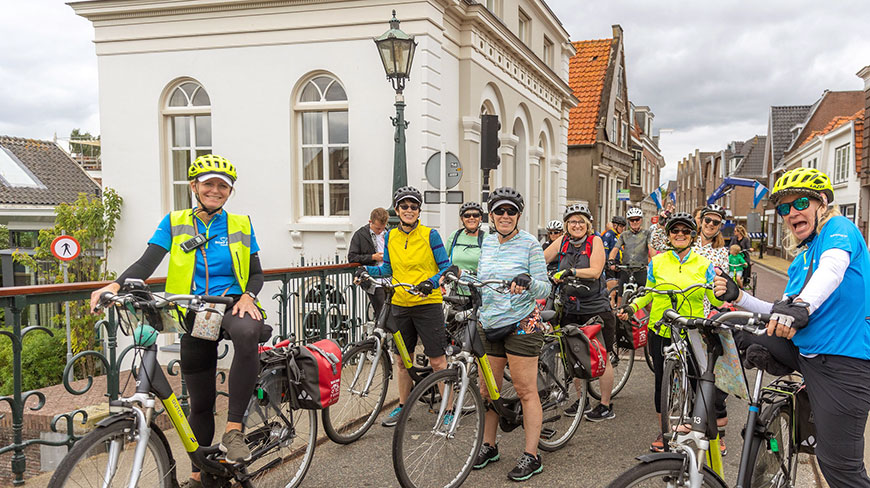 A group of smiling cyclists in helmets pose with their bikes on a charming street in the Netherlands.