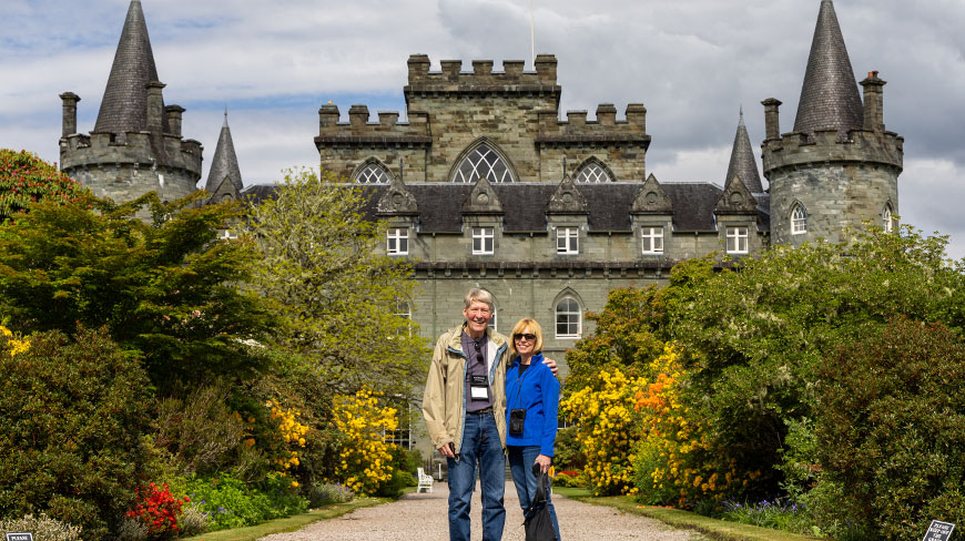 A couple smiles for a photo on a path in the lush gardens of Inveraray Castle in Scotland.