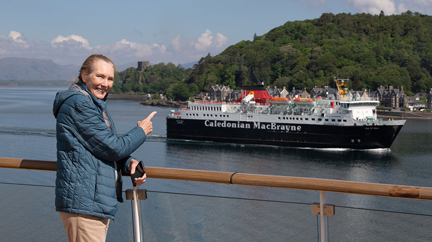 A woman on a ship in Scotland points at a Caledonian MacBrayne ferry with a green coastline behind it.