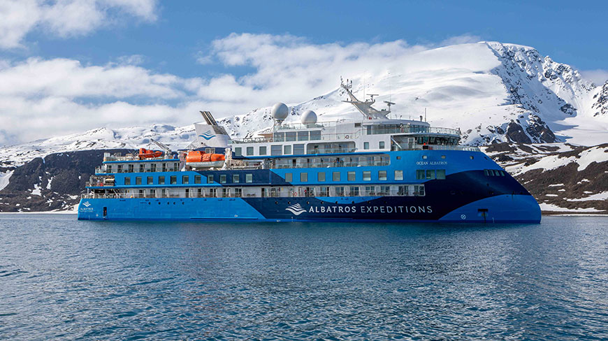 The Ocean Albatros expedition ship floats in the calm waters of Antarctica, with a massive snow-covered mountain range rising in the background.
