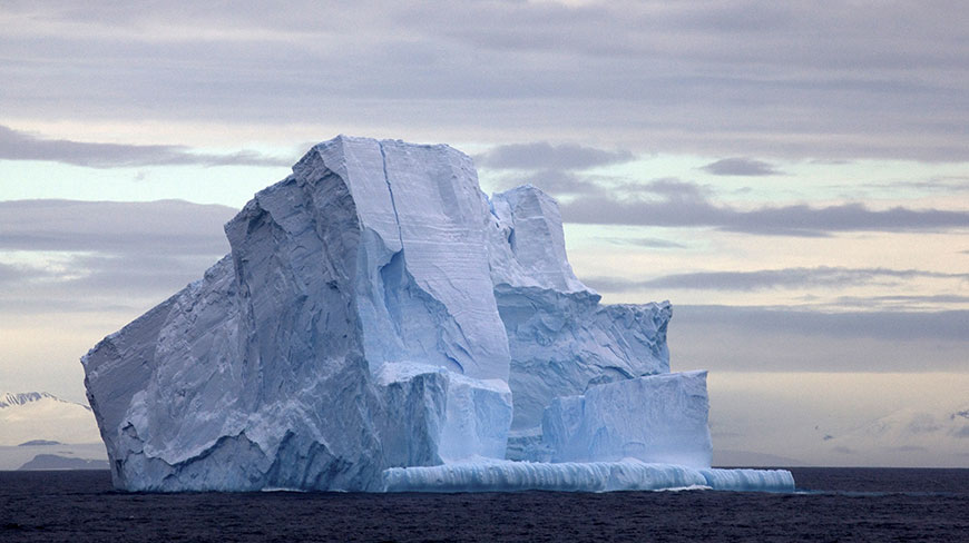 A large, jagged iceberg floats in the dark waters of the Drake Passage in Antarctica under a cloudy, grey sky.