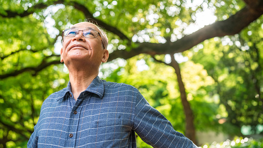An older Asian man wearing glasses and a blue plaid shirt looks up peacefully while standing outside under green trees.