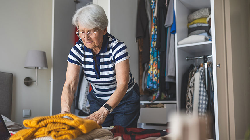 An older woman with white hair and glasses folds sweaters on a bed in her bedroom with the closet door open behind her.