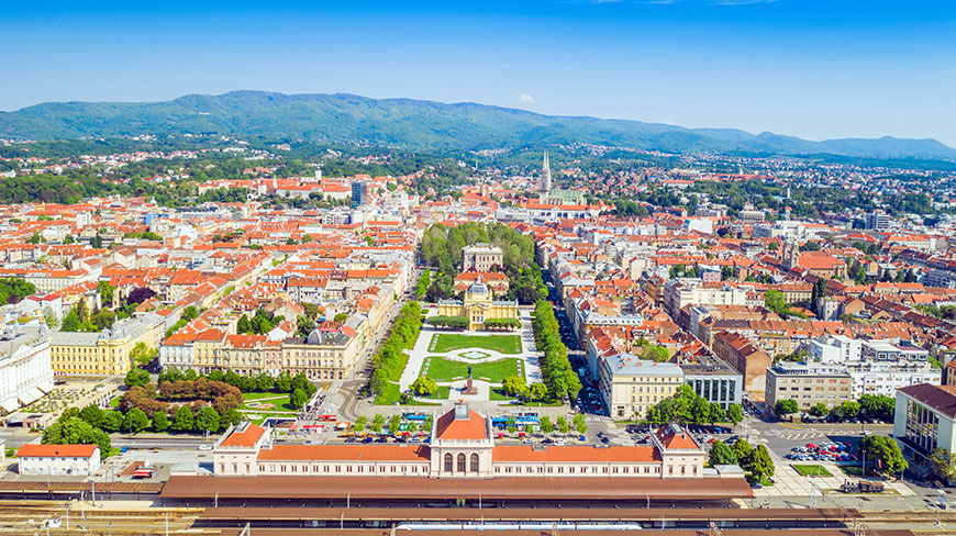 Aerial view of Zagreb, Croatia, showing the city's red-roofed buildings, the central Green Horseshoe park, and mountains in the distance.