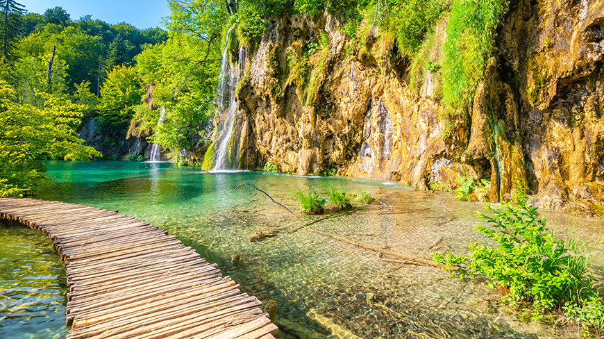 A wooden boardwalk curves over clear turquoise water towards waterfalls at Plitvice Lakes National Park in Croatia.