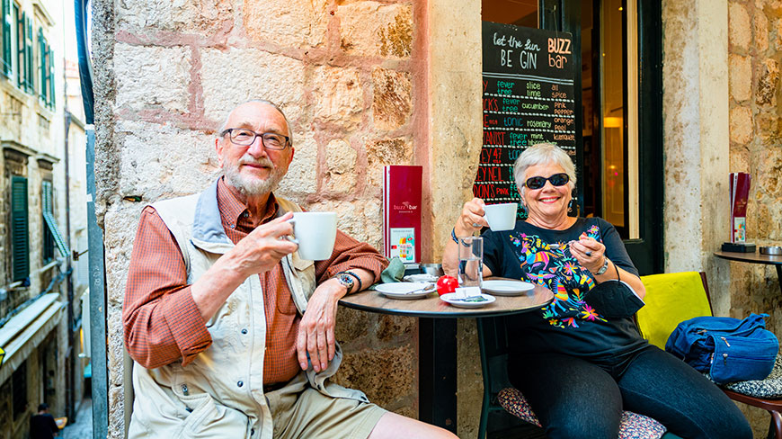 An older couple smiles while enjoying coffee at an outdoor cafe on a narrow stone street in Dubrovnik, Croatia.