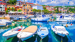 Several boats are moored in the bright blue harbor of Opatija, Croatia, with colorful hillside buildings of the coastal town in the background.