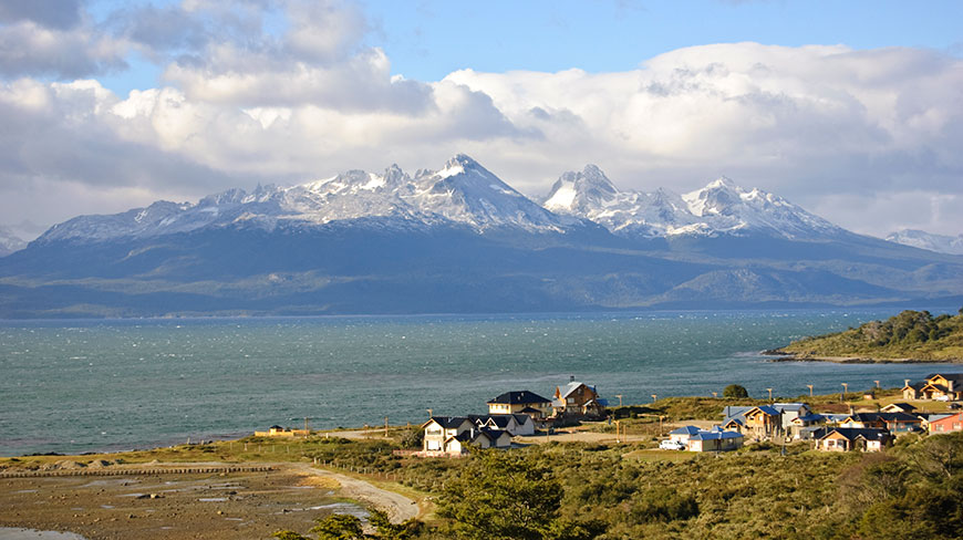 A small coastal town in Argentina sits on the Beagle Channel, with jagged, snow-capped mountains rising in the distance under a cloudy sky.