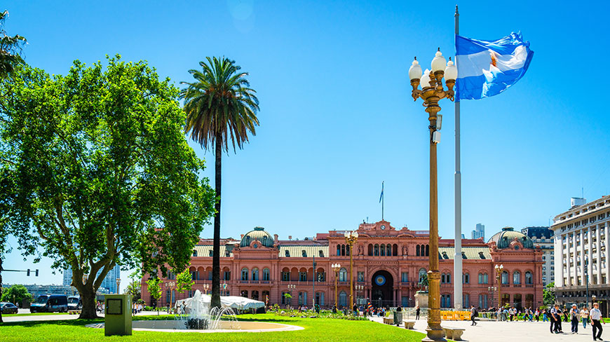 The pink Casa Rosada in Buenos Aires, Argentina, is seen from a park with an Argentinian flag waving under a clear blue sky.