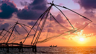 Chinese fishing nets in Kochi are silhouetted against a dramatic purple and gold sunset over the ocean.