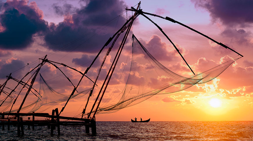 Large, traditional Chinese fishing nets are silhouetted against a vibrant purple and orange sunset over the water in Kochi, Kerala.