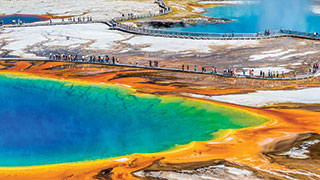 An aerial view of the Grand Prismatic Spring in Yellowstone National Park, with tourists walking on a boardwalk along its vibrant, colorful edge.