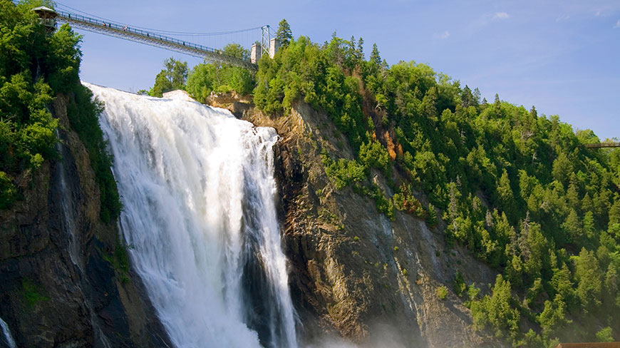 Montmorency Falls in Quebec, Canada, plunges over a cliff, with a pedestrian bridge crossing above the lush, green forest.