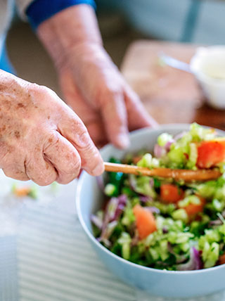 An older person's hands use a wooden spoon to mix a colorful salad in a light blue bowl.
