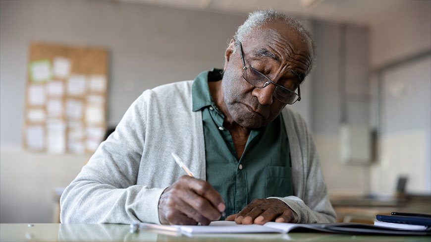 An older Black man wearing glasses and a cardigan sits at a desk, writing on a piece of paper with a pen.