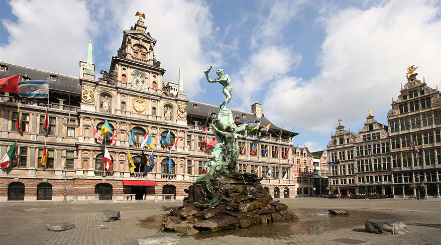 The Grote Markt in Antwerp, Belgium, featuring the historic City Hall decorated with flags and the central Brabo Fountain under a partly cloudy sky.