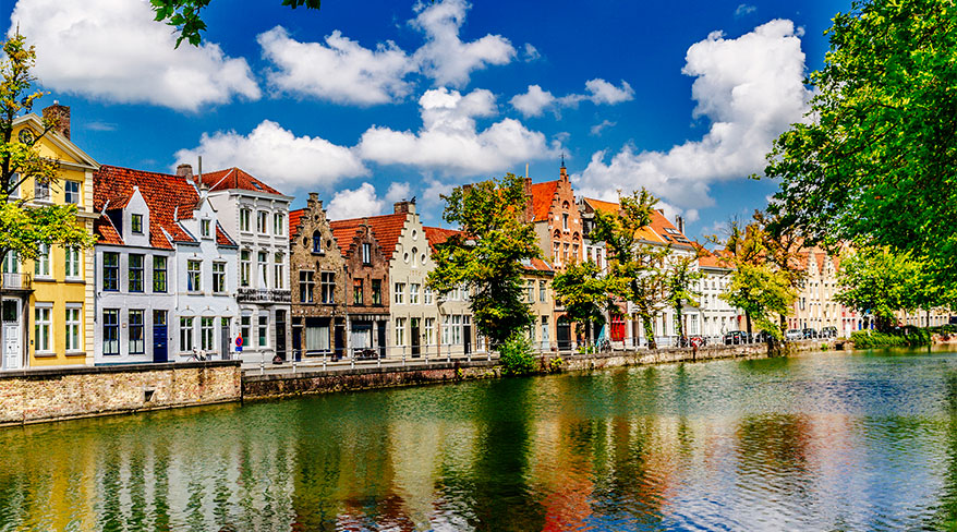 Colorful, historic buildings line a canal in Bruges, Belgium, under a bright blue sky with fluffy white clouds.