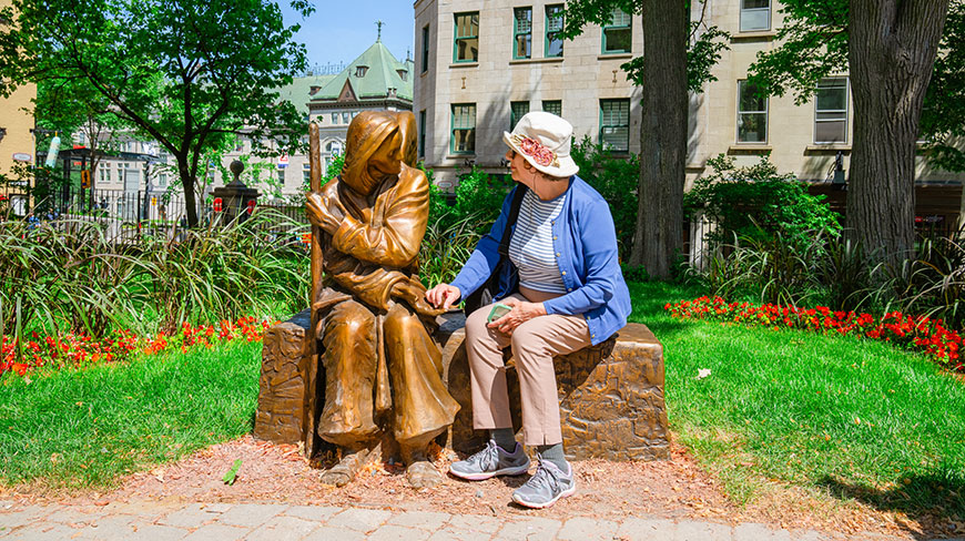 A woman sits on a bench in a park in Quebec City, Canada, interacting with a bronze statue of a seated figure.