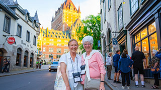 Two women smile for the camera on a cobblestone street in Old Quebec, Canada, with the grand Château Frontenac hotel in the background.