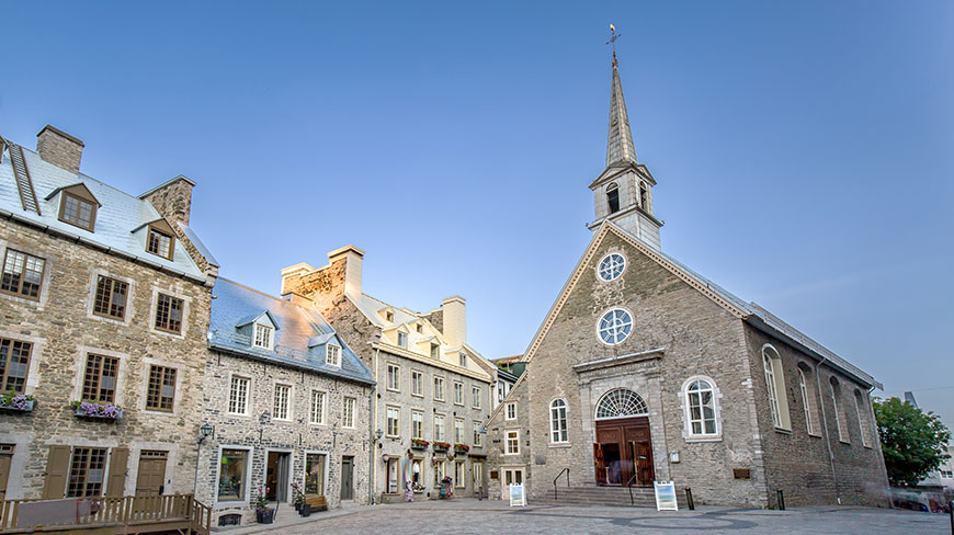 The historic stone Notre-Dame-des-Victoires Church stands in a public square in Québec City, Canada, surrounded by old buildings under a clear sky.