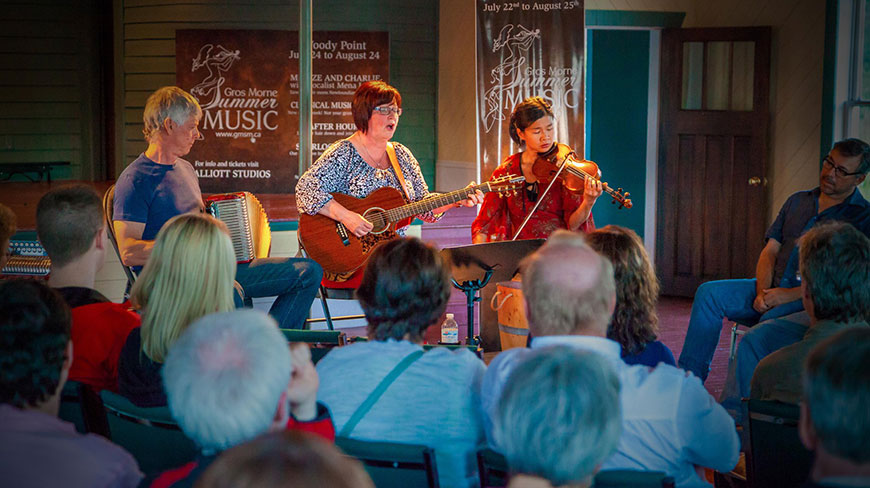 Musicians perform with a guitar, violin, and accordion for an audience at the Gros Morne Musical Theatre in Newfoundland and Labrador.