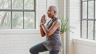 An older woman with short gray hair stands in a yoga tree pose in a brightly lit room with large windows.