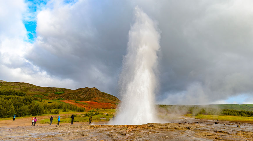 A large geyser erupts in a dramatic display for a group of onlookers against a backdrop of rolling green hills in Iceland.