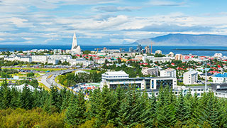 A scenic view of Reykjavik, Iceland, with the iconic Hallgrímskirkja church, the sea, and mountains in the distance, framed by green trees.