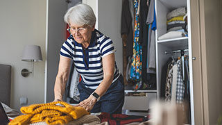 An older woman with white hair folds a yellow sweater in her bedroom next to an open closet.